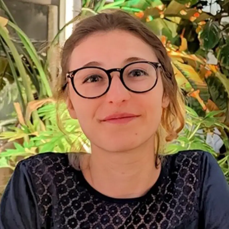 A person with glasses and light brown hair smiles slightly whilst seated indoors, with green plants in the background and wearing a dark-patterned top.