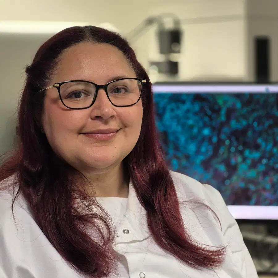 A woman with long dark red hair and glasses wears a white lab coat and smiles whilst sitting in front of a computer showing a blue and black scientific image.