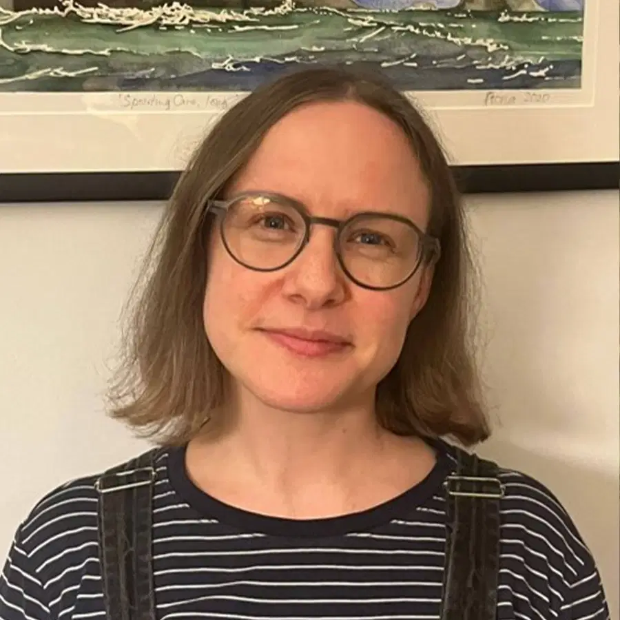 A person with shoulder-length light brown hair and glasses smiles softly, wearing a striped shirt and dark dungarees, standing in front of a framed painting of waves.