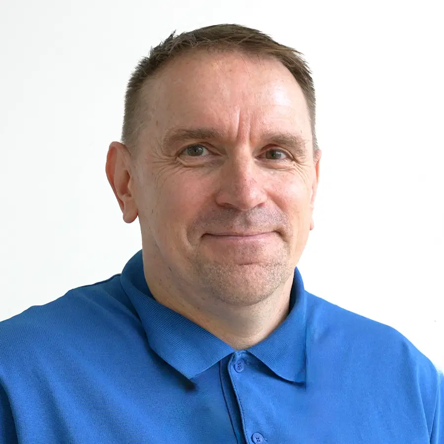 A middle-aged man with short brown hair smiles softly. He is wearing a bright blue collared shirt and stands in front of a plain white background.