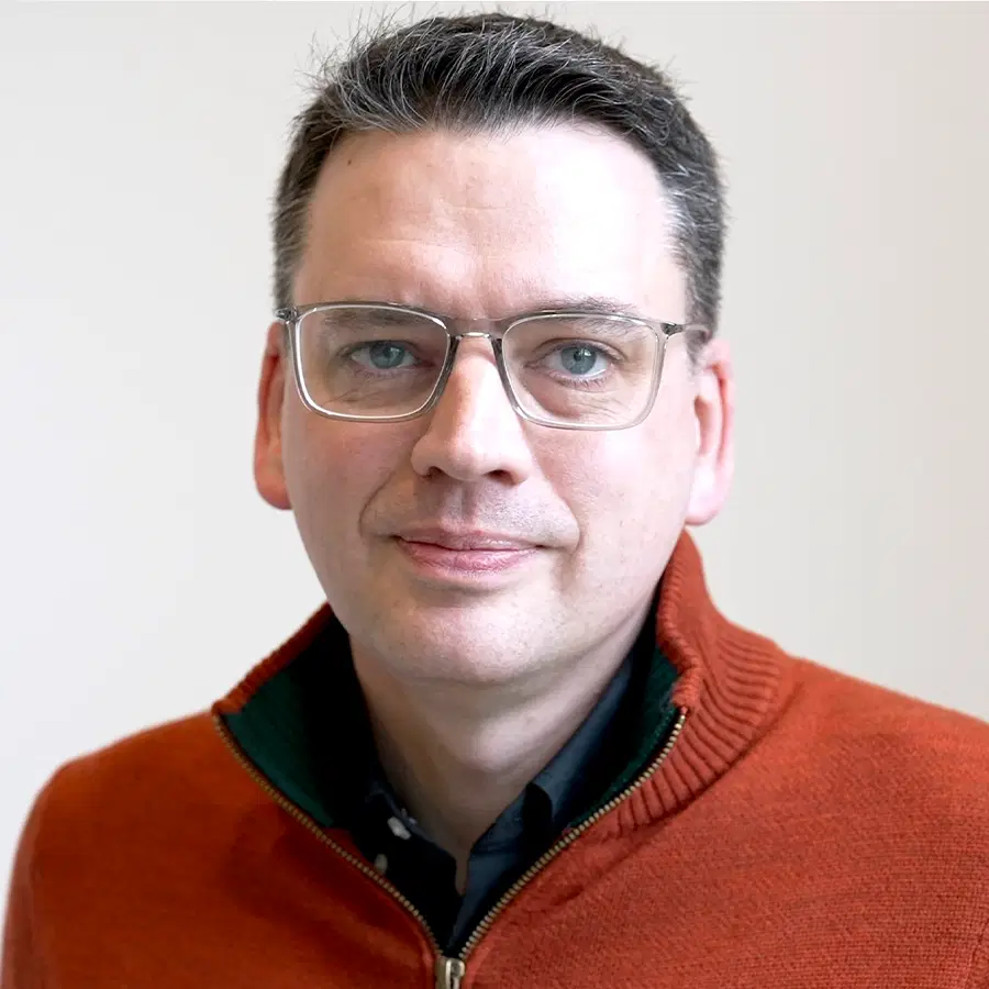A man with short dark hair, wearing glasses, a dark shirt, and a red zip-up jumper, looks at the camera against a plain light background.