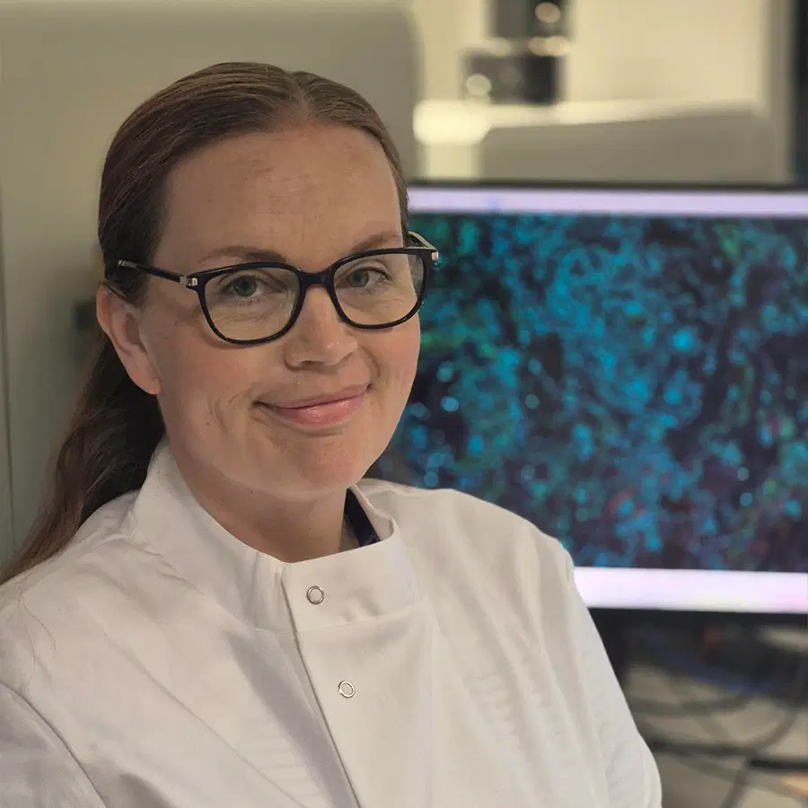 A woman wearing glasses and a white lab coat smiles in a laboratory setting, with a computer monitor displaying a blue and black image in the background.