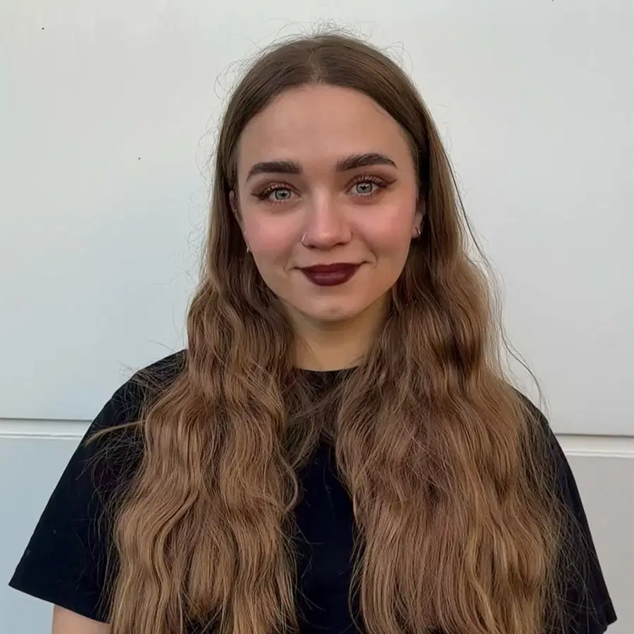 A woman with long wavy brown hair and brown lipstick stands in front of a plain light background, wearing a black shirt and smiling softly at the camera.