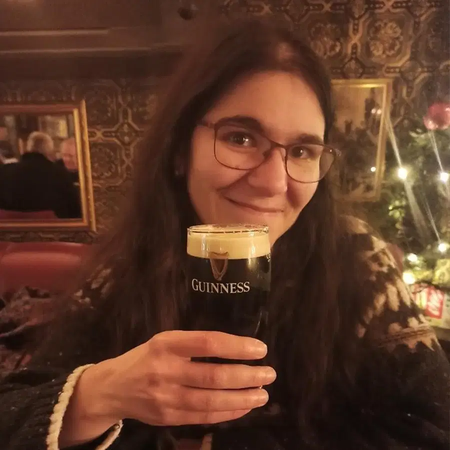 A woman with long dark hair and glasses smiles whilst holding a pint of Guinness in a cosy pub with patterned wallpaper and warm lighting.