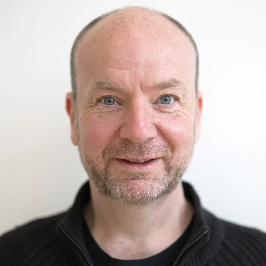 A middle-aged man with a beard and short hair smiles slightly at the camera, wearing a dark zip-up jumper against a plain white background.