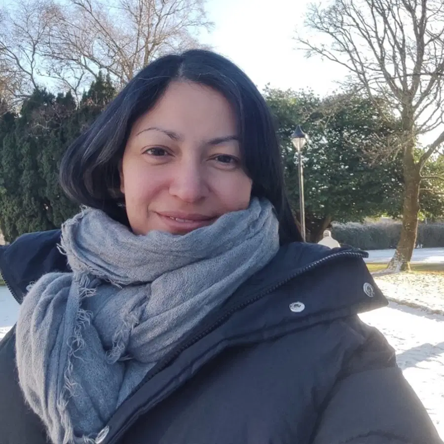 Woman with long dark hair in a grey scarf and black coat smiles outdoors on a sunny winter’s day, with snow on the ground and trees in the background.