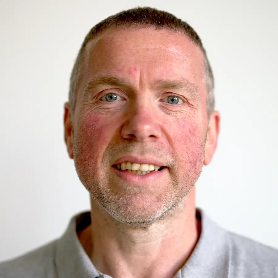 A middle-aged man with short hair, light eyes, and a trimmed beard, wearing a light grey polo shirt, is smiling slightly against a plain white background.
