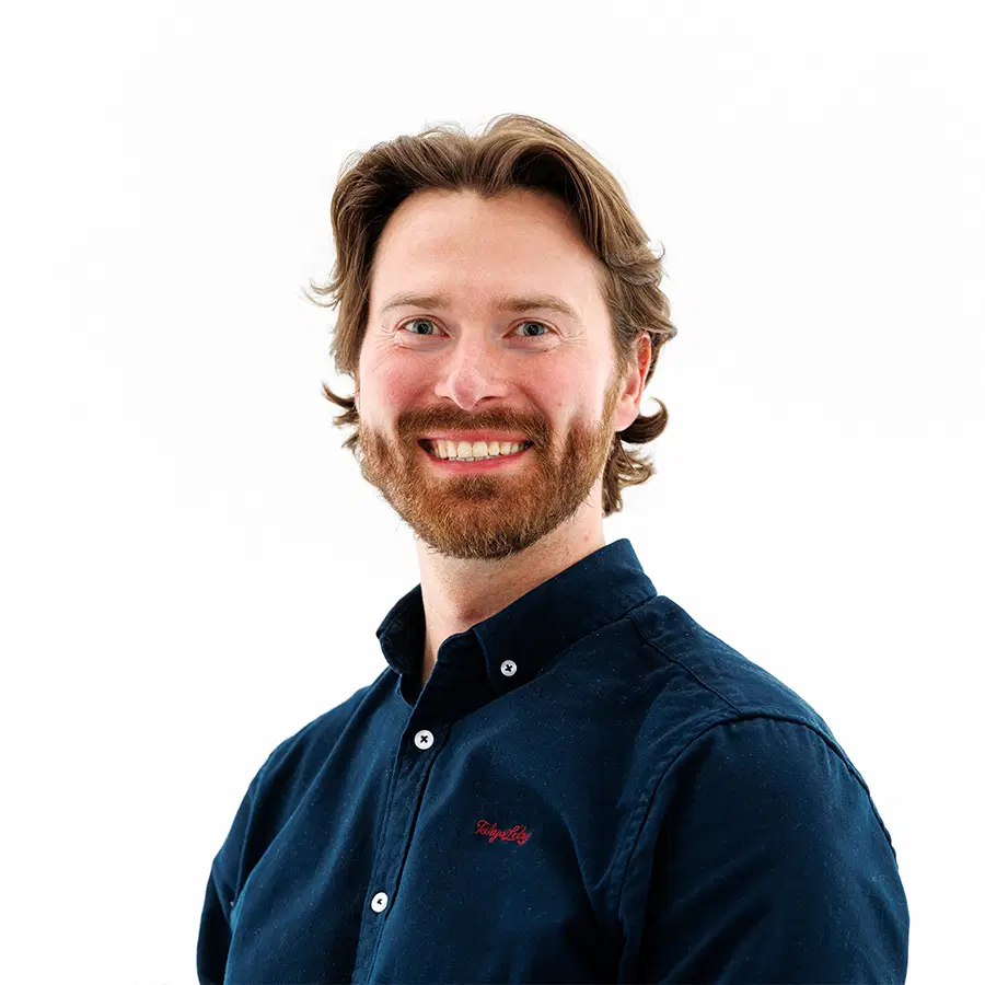 A smiling man with light skin, brown hair, and a beard is wearing a navy blue button-up shirt. He is standing in front of a plain white background.