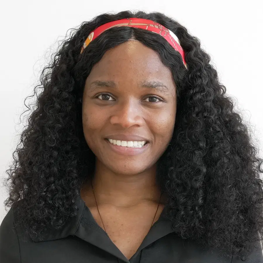 A woman with curly black hair and a red headband smiles at the camera. She is wearing a black collared shirt and a necklace, and is posed against a plain white background.