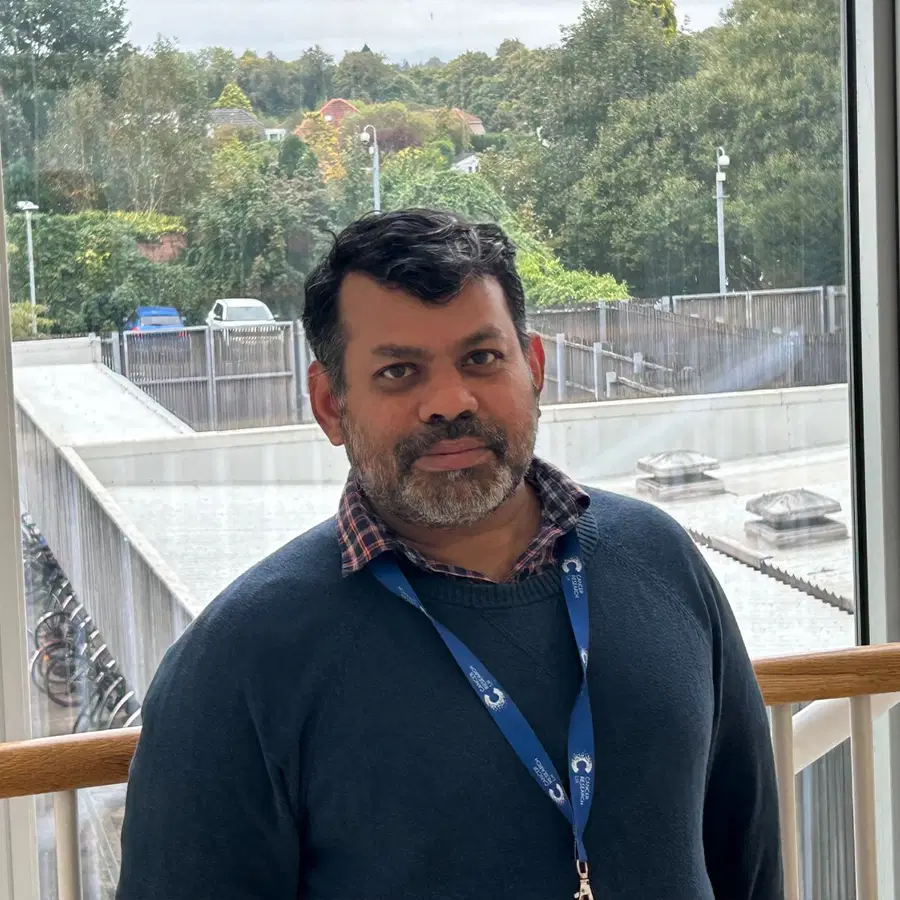 A man with a beard and dark hair, wearing a blue jumper and checked shirt, stands indoors by a window with a lanyard, with trees and buildings visible outside.