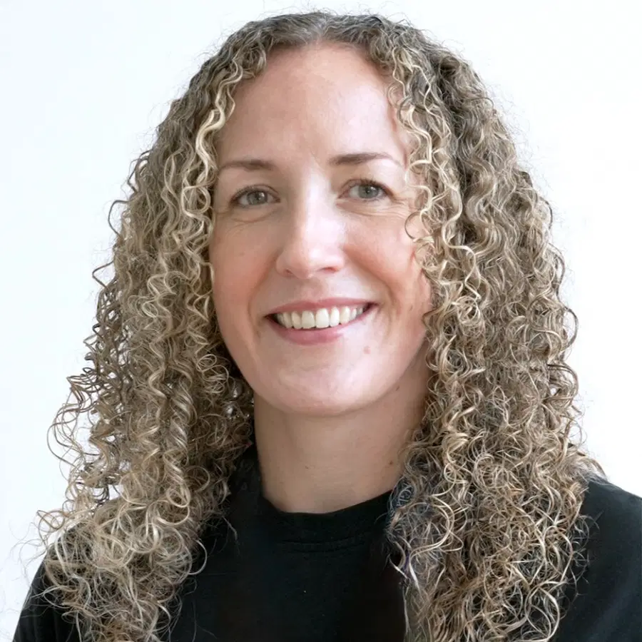 A person with long, curly, light brown hair and blue eyes smiles at the camera. They are wearing a black top and standing in front of a plain white background.