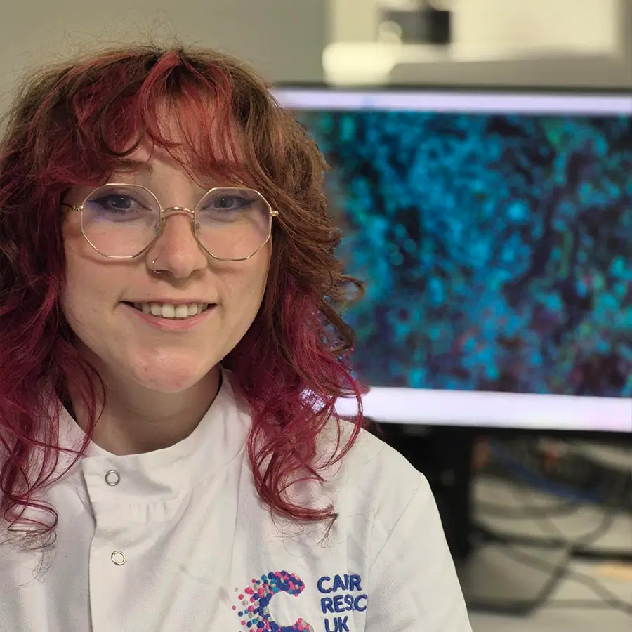 A person with curly red hair and glasses smiles, wearing a white lab coat in front of a computer monitor displaying a blue and black scientific image.