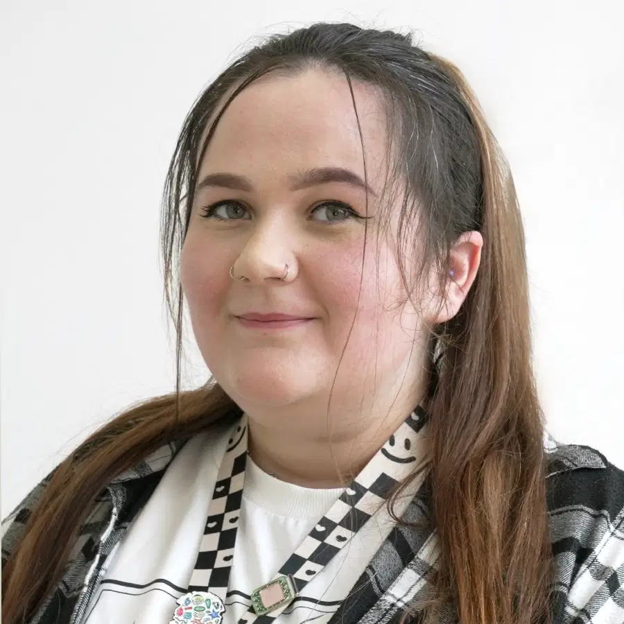 A woman with long brown hair in a ponytail, wearing a checked shirt, white graphic T-shirt, and chequered lanyard, smiles softly at the camera against a plain white background.