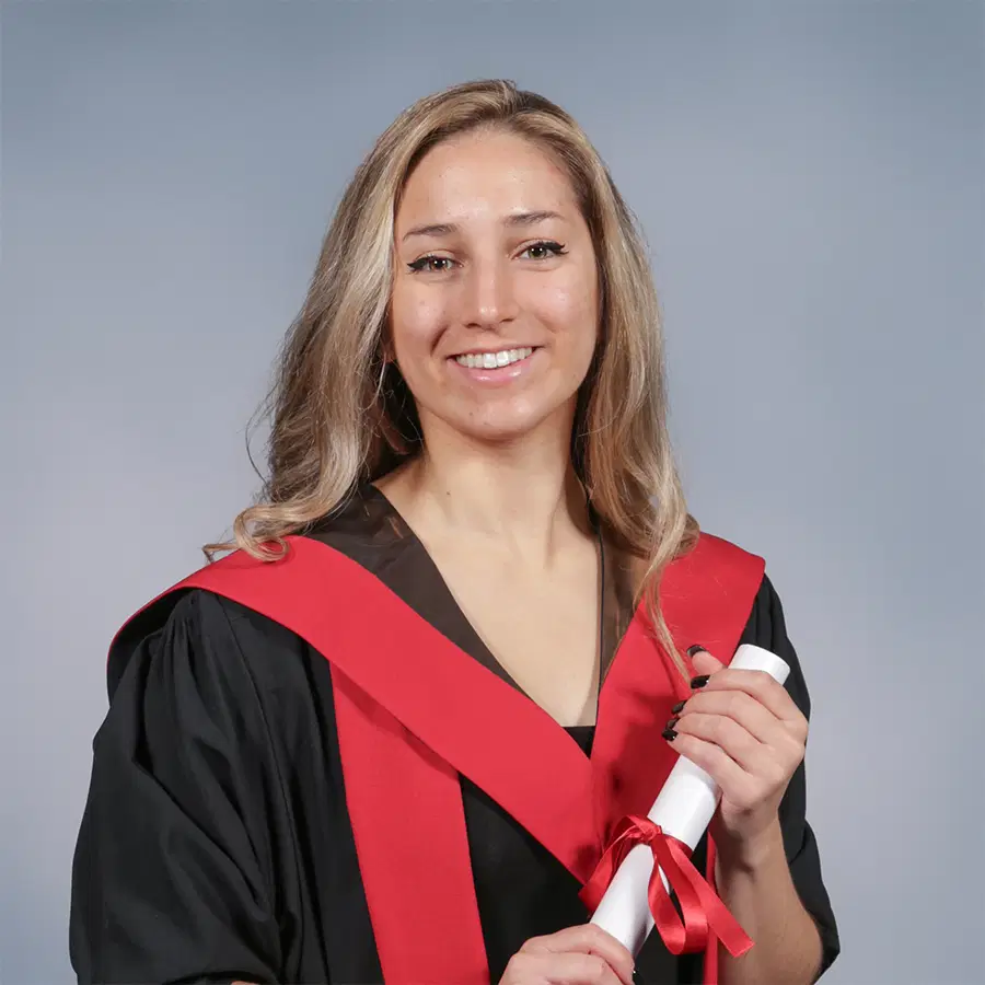 A woman in a black graduation gown with a red sash holds a rolled-up diploma, smiling at the camera against a plain light blue background.