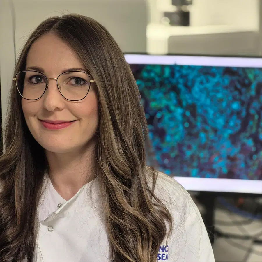 A woman with long brown hair and glasses is smiling, wearing a white lab coat. She is standing in front of a computer screen displaying a colourful scientific image.