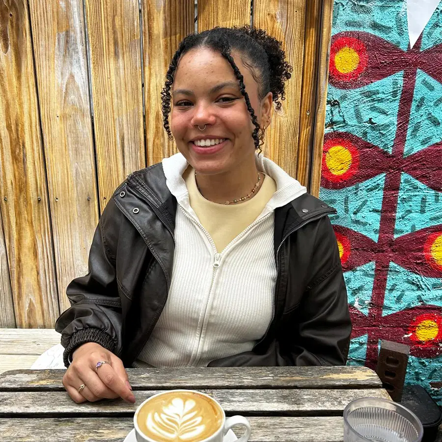 A person sits at an outdoor table with a latte, smiling at the camera. Behind them is a wooden fence and a colourful mural featuring red circles and blue patterns.
