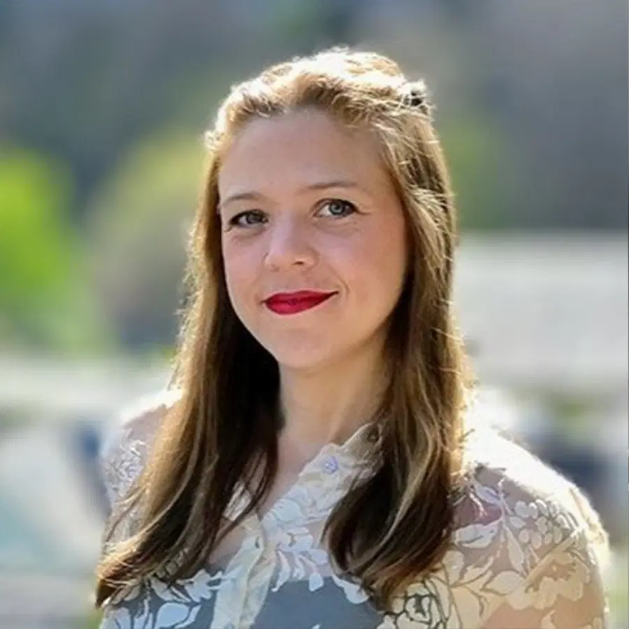 A woman with long brown hair and red lipstick smiles outdoors, wearing a floral-patterned top. The background is blurred with greenery and soft light.