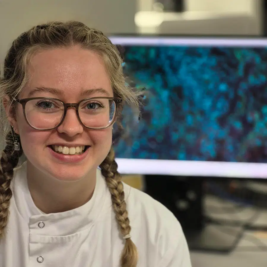 A smiling person with glasses and plaited hair, wearing a white lab coat, sits in front of a computer displaying a blue and black scientific image.