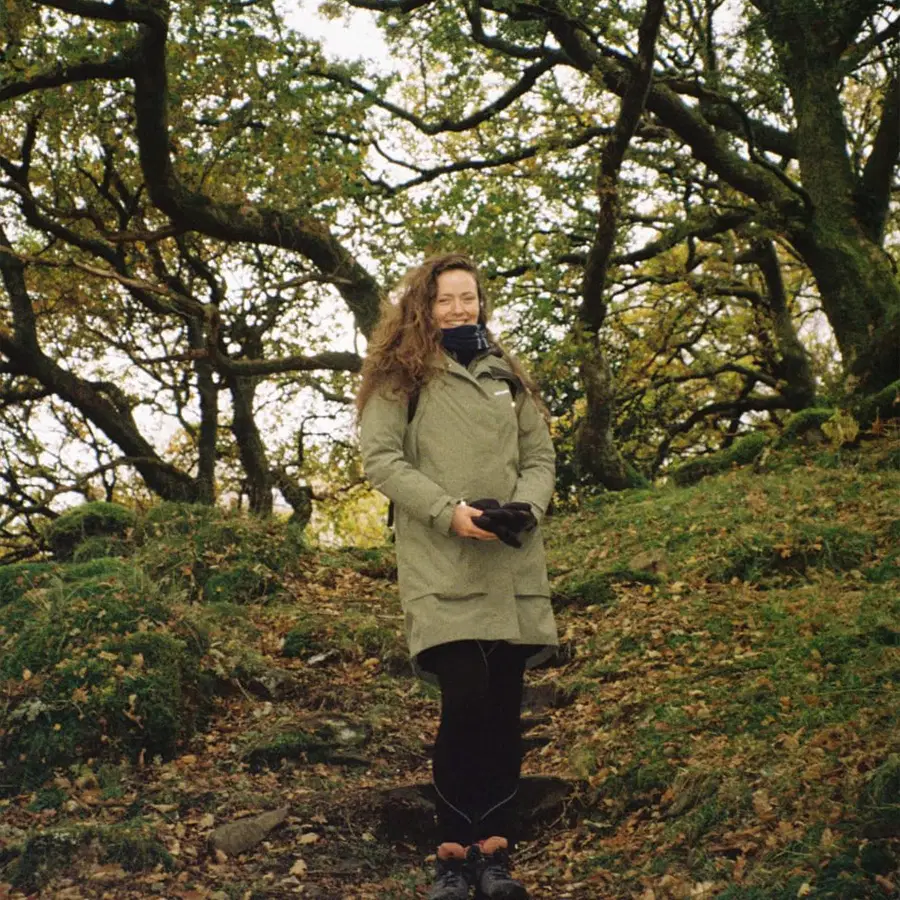A woman with curly hair stands on a forest path, wearing a light green coat and scarf. She holds gloves and smiles with her eyes closed, surrounded by green trees and fallen leaves.