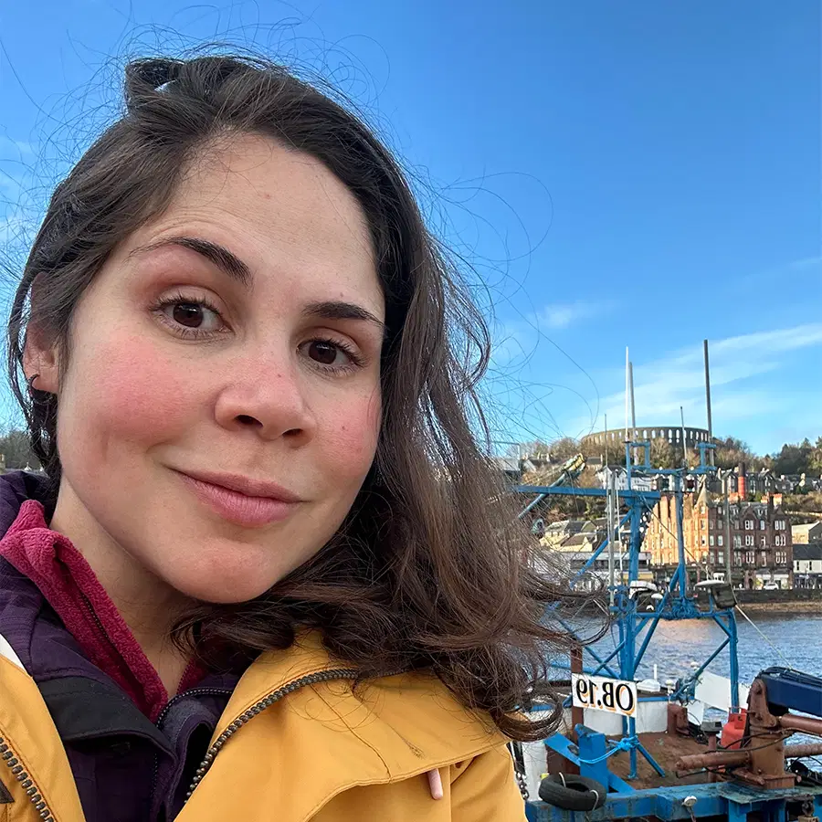 A woman in a yellow jacket takes a selfie by a harbour with blue sky, boats, water, and buildings in the background.