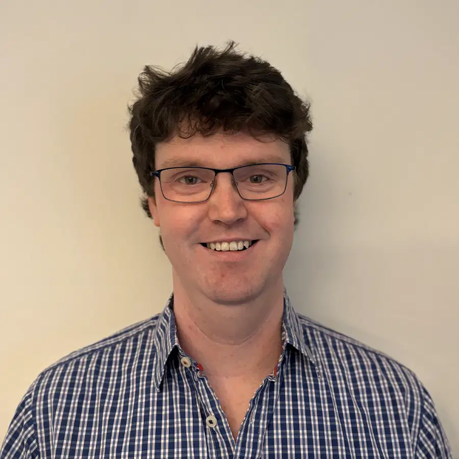 A person with short, curly brown hair and glasses is smiling at the camera. They are wearing a blue and white checked shirt and are posed in front of a plain, light-coloured background.