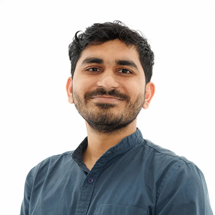 A man with short, wavy dark hair and a beard is smiling slightly. He is wearing a blue button-up shirt and is standing against a plain white background.