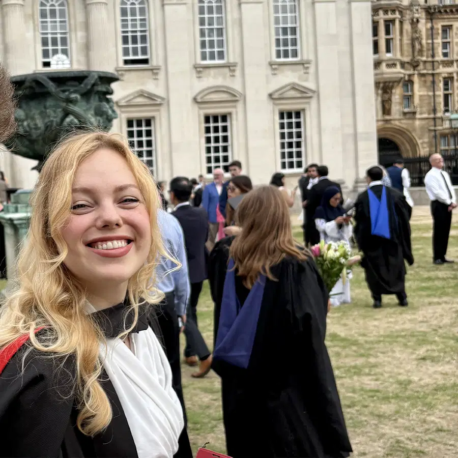 A smiling graduate in a cap and gown stands outdoors among other graduates and guests in front of historic university buildings.