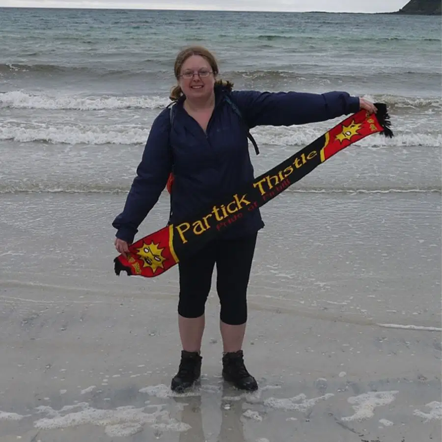 A smiling person stands on a beach with waves behind them, holding a red and yellow Partick Thistle scarf. They wear glasses, a dark jacket, cropped trousers, and boots.