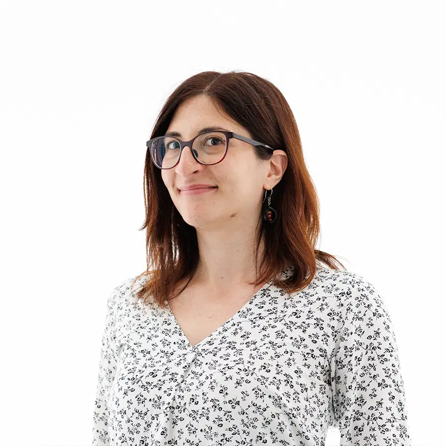 A woman with shoulder-length brown hair, glasses, and a patterned white blouse smiles slightly whilst looking at the camera against a plain white background.