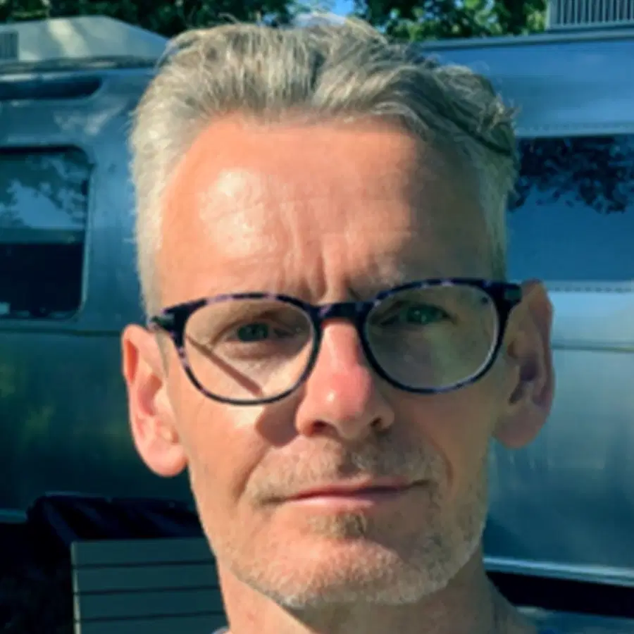 A man with short grey hair and black-rimmed glasses stands outside in sunlight, with a metallic caravan and trees in the background.
