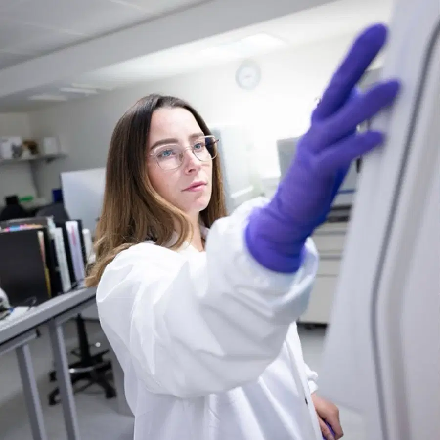 A person in a white lab coat and purple gloves interacts with laboratory equipment in a bright laboratory. Shelves and office equipment are visible in the background.