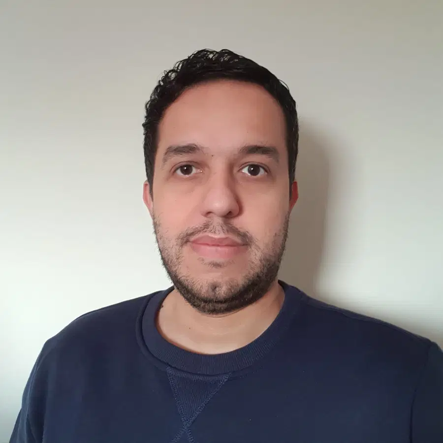 A man with short, dark curly hair and a trimmed beard is wearing a navy blue jumper and standing against a plain, light-coloured wall.