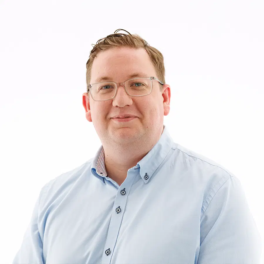 A man with short brown hair and glasses smiles slightly whilst wearing a light blue button-up shirt against a plain white background.