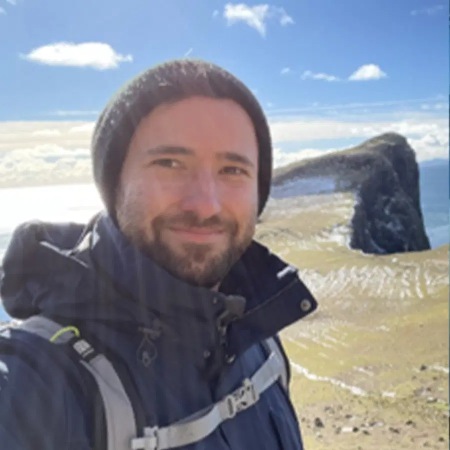 A person wearing a winter jacket and beanie smiles at the camera with a rocky coastline, blue sky, and sea in the sunny background.