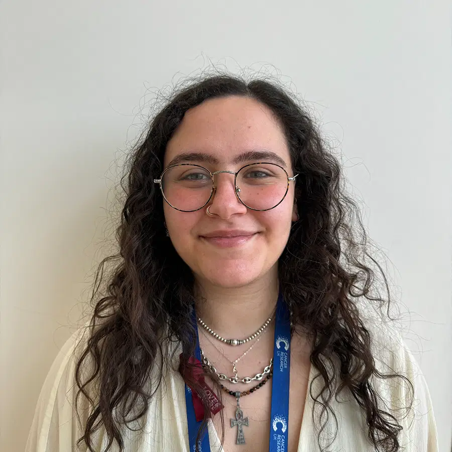 A person with long curly hair and glasses smiles at the camera. They wear a white shirt, layered necklaces, and a blue lanyard. The background is a plain, light-coloured wall.