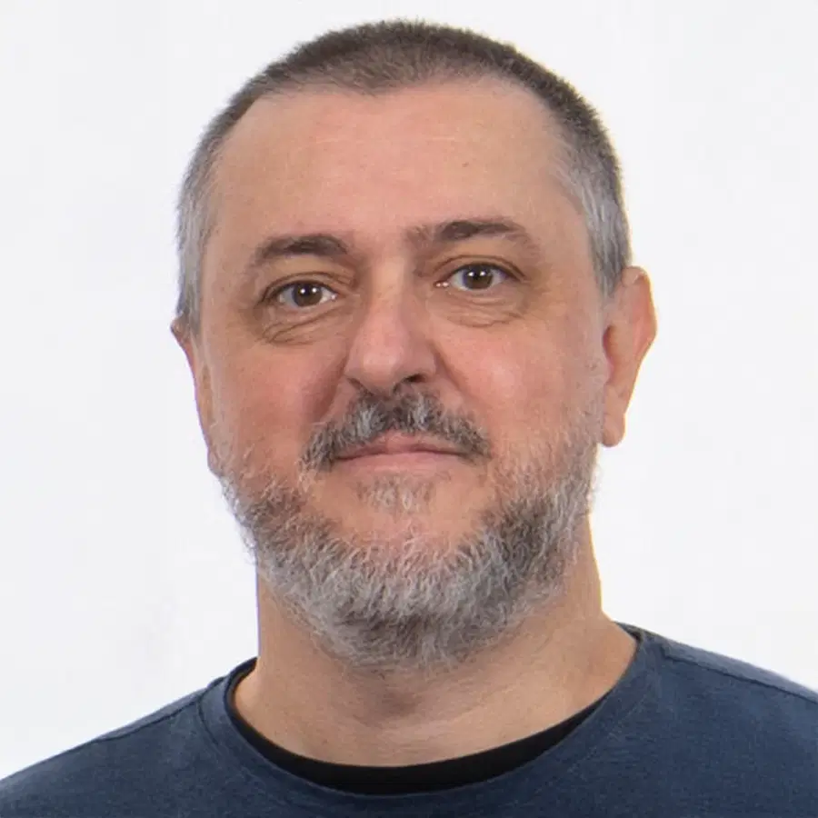 A middle-aged man with short grey hair and a beard, wearing a blue crew-neck jumper, is smiling slightly in front of a plain white background.