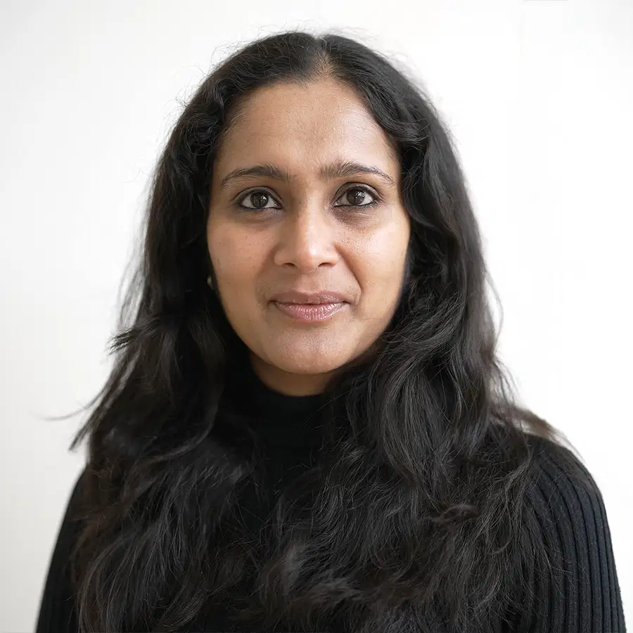 A woman with long, dark hair and medium skin tone, wearing a black polo neck jumper, looks at the camera against a plain white background.