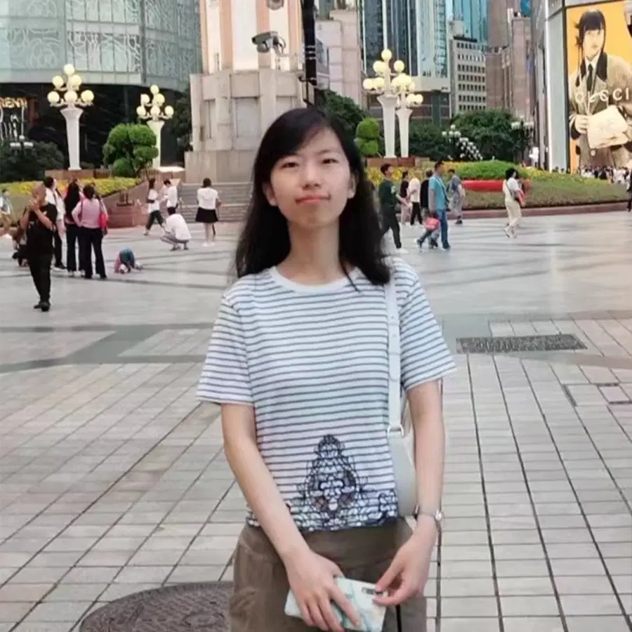 A young woman in a striped shirt stands in a busy city square with people walking in the background, surrounded by buildings, greenery, and streetlights.