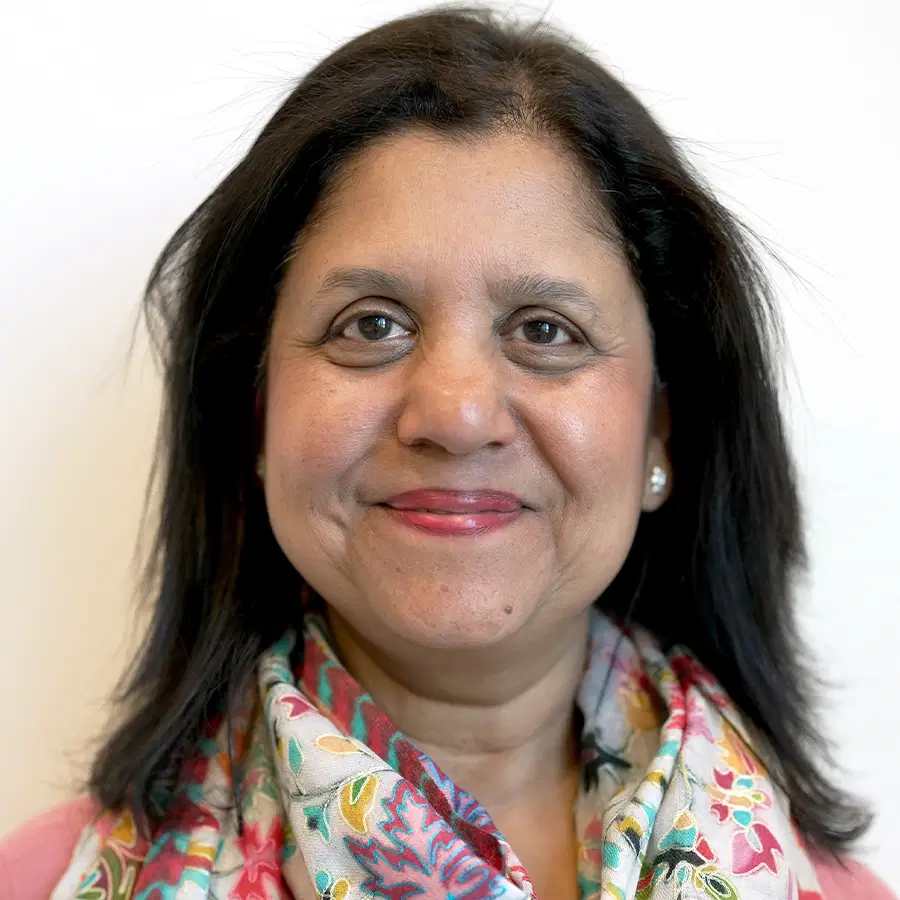 A woman with medium-length dark hair smiles at the camera. She wears a colourful floral scarf and a pink top, with a light background behind her.