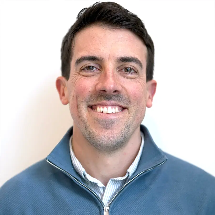 A man with short dark hair smiles at the camera, wearing a light blue zip-up jumper over a collared shirt, standing in front of a plain white background.