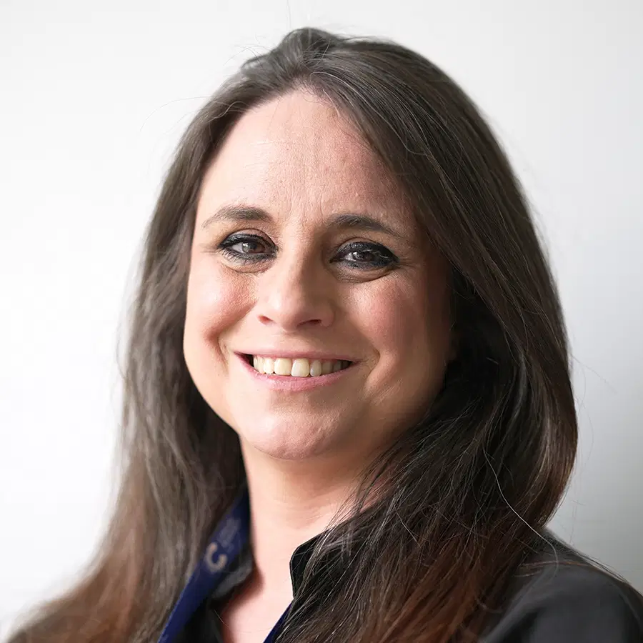 A woman with long brown hair and a black top smiles at the camera in front of a plain white background.