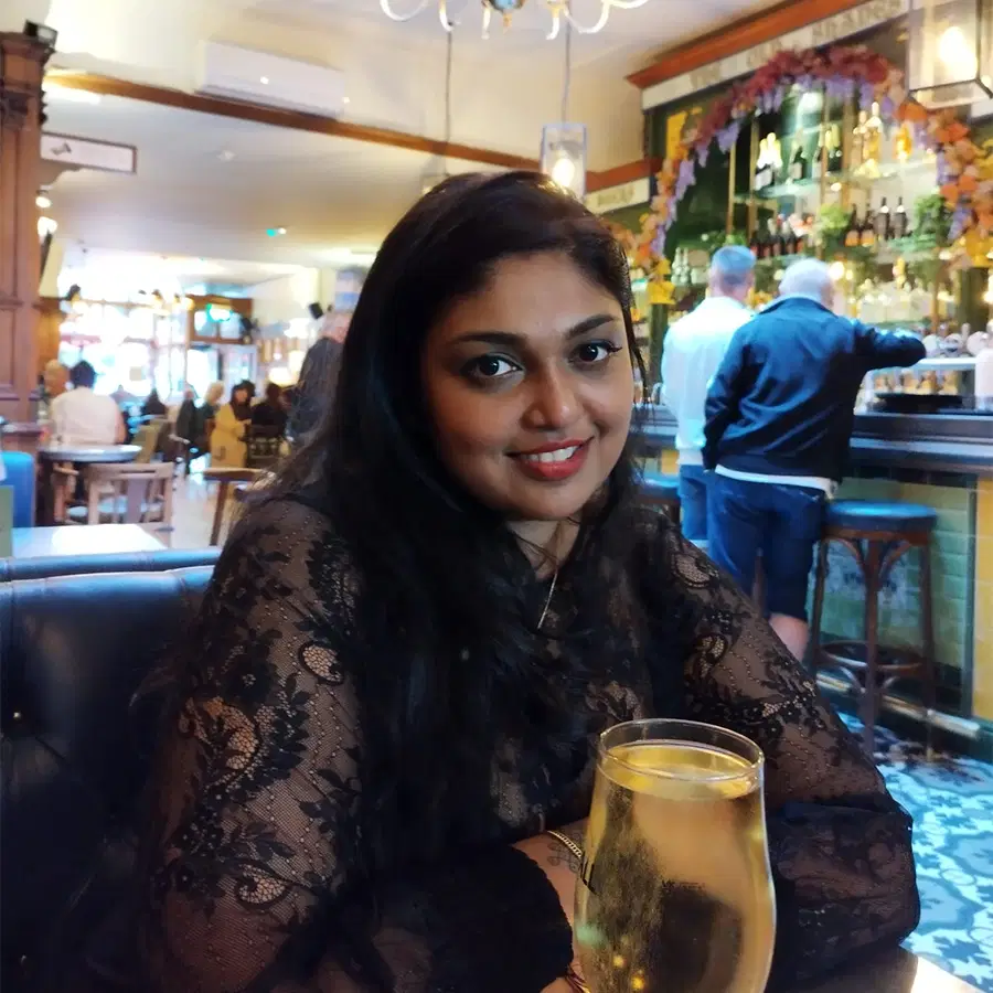 A woman with long dark hair, wearing a black lace top, sits at a bar holding a glass of white wine. She is smiling at the camera, with people and shelves of bottles visible in the background.