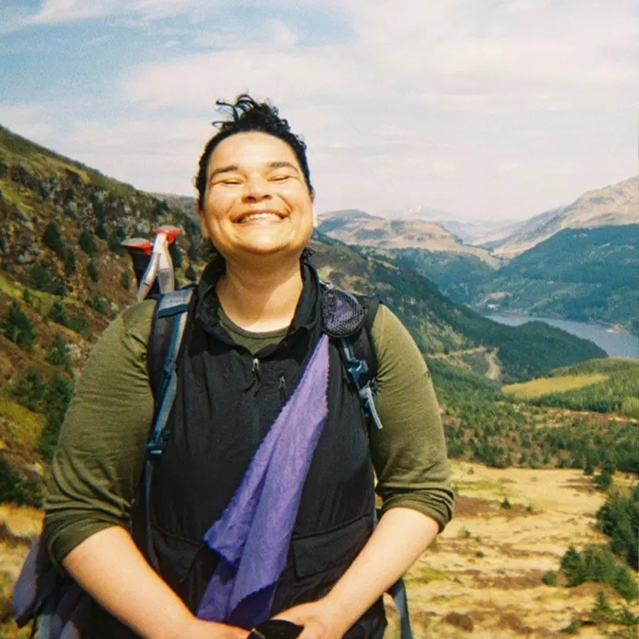 A person with dark hair smiles broadly while walking in a mountainous area with green hills and a lake in the background. The weather is clear and sunny.