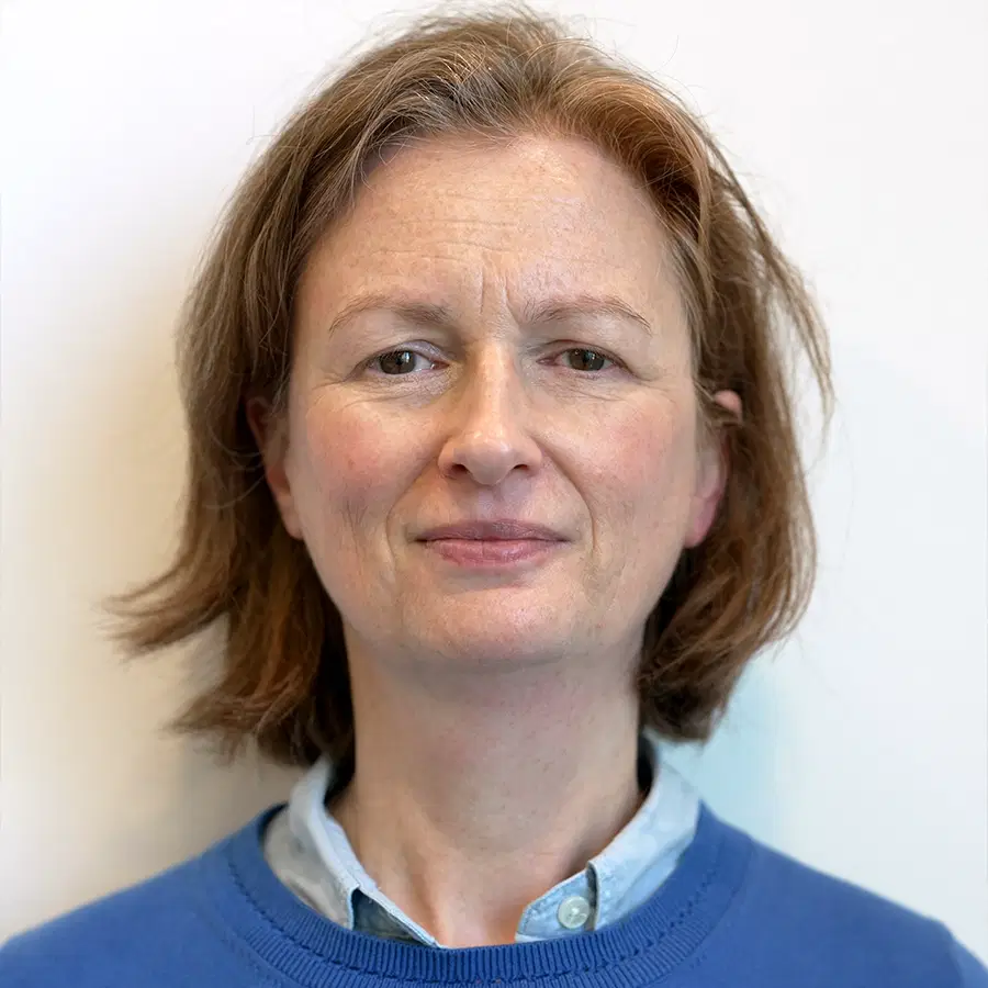 A middle-aged person with short brown hair, wearing a blue jumper over a collared shirt, stands against a plain white background, looking directly at the camera with a neutral expression.