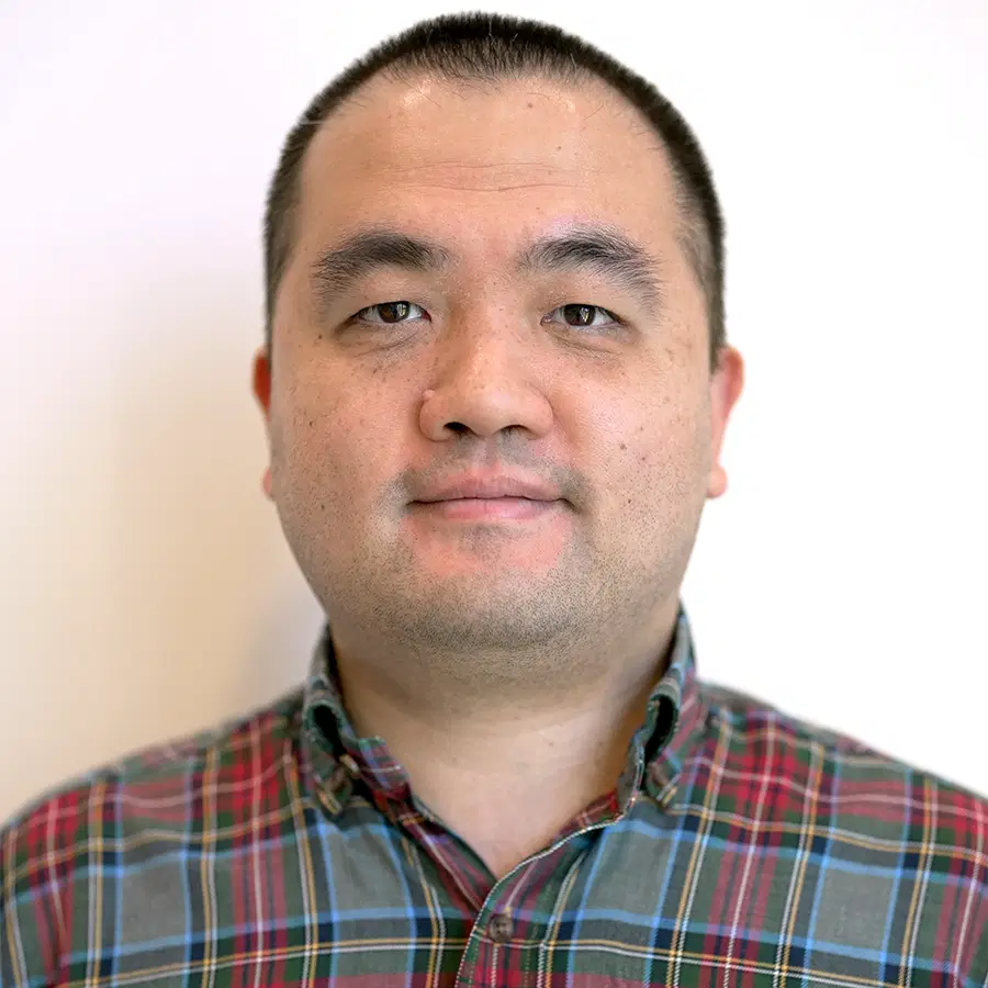 A man with short dark hair and a trimmed beard is wearing a red, blue, and green checked button-up shirt, standing against a plain light background.