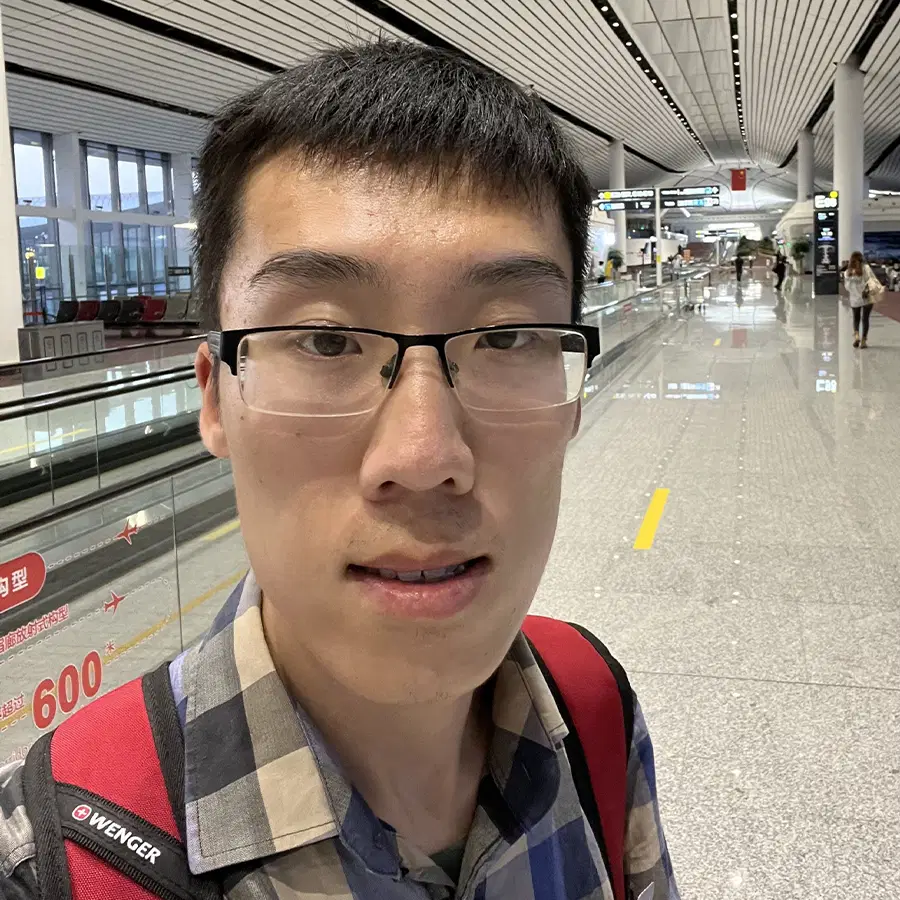 A person wearing glasses and a red rucksack takes a selfie in a spacious, modern airport terminal with shiny floors, high ceilings, and glass walls.