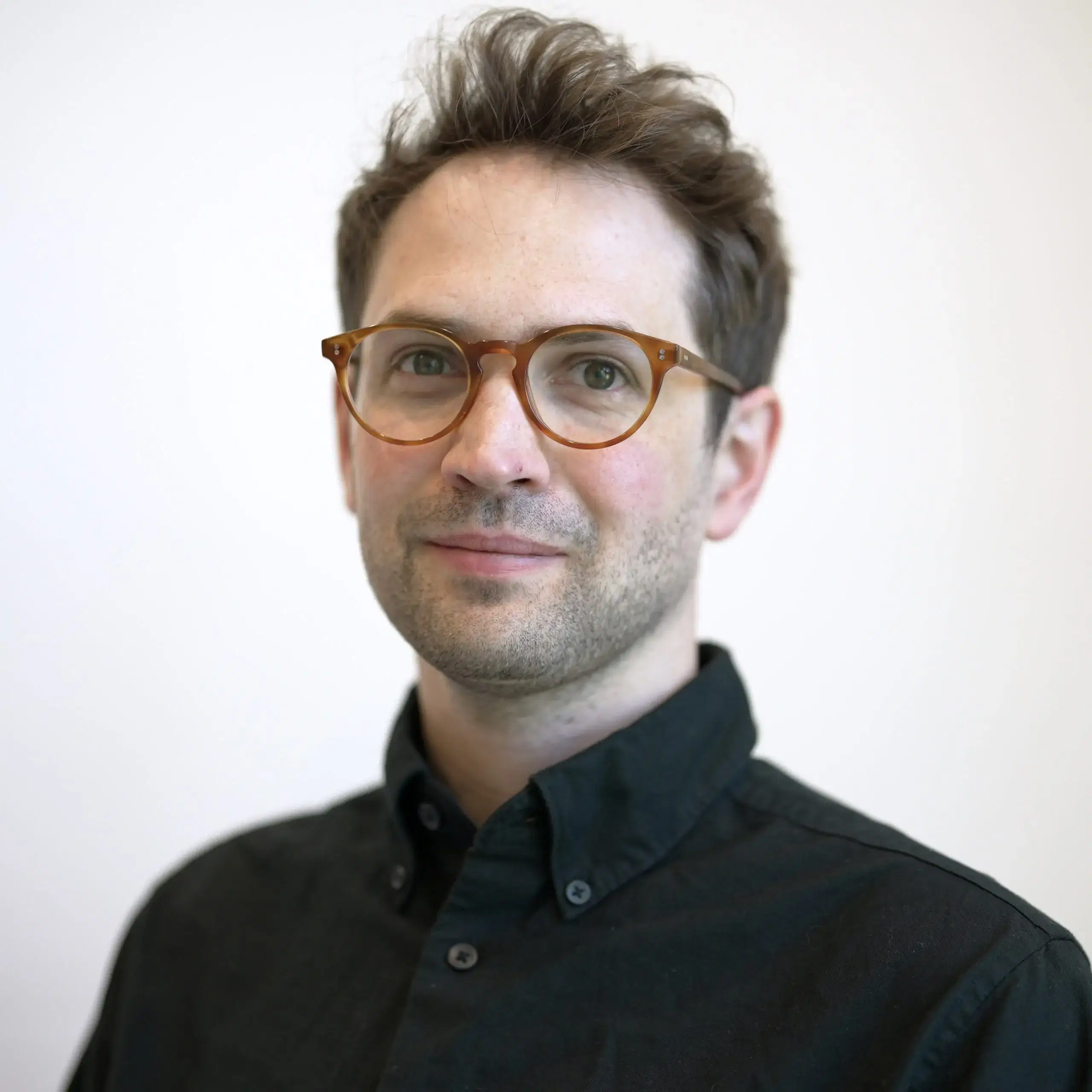 A man with short brown hair, light skin, and brown glasses wears a black button-up shirt and looks at the camera, standing against a plain light background.
