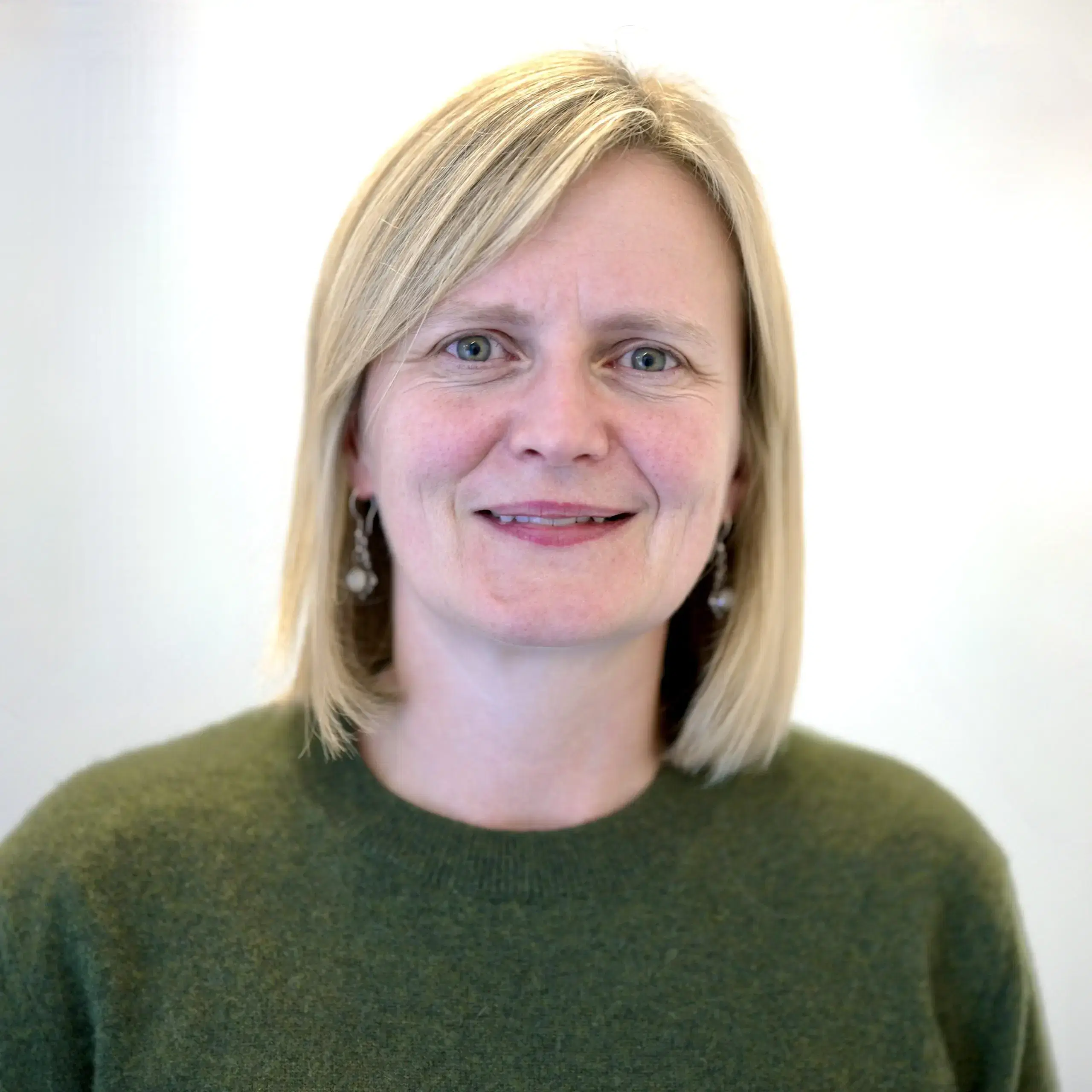 A woman with straight blonde hair, wearing a green jumper and silver earrings, smiles gently at the camera against a plain light background.