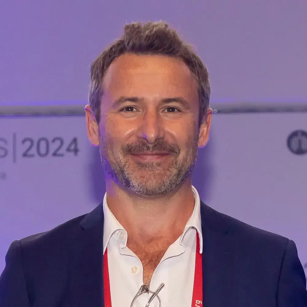 A man with short brown hair and a beard smiles at the camera, wearing a suit jacket and an open-collared white shirt. A conference backdrop with 2024 is visible behind him.