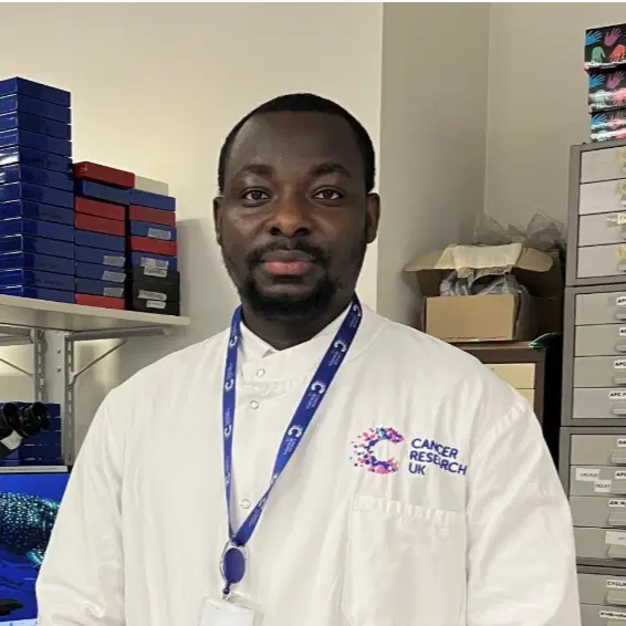 A person in a white Cancer Research UK lab coat and lanyard stands in a laboratory with shelves, boxes, and filing cabinets in the background.